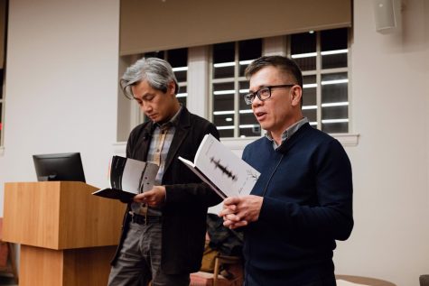 A man with grey hair, a plaid button-down, and a brown jacket appears to read from a book of poetry. To his right, a man with black hair, rectangular black glasses, and a blue v-neck sweater over a grey button-down is holding a book and is mid-speech. Both men are standing to the side of a wooden podium. A large, dark window is visible on the white wall behind them.