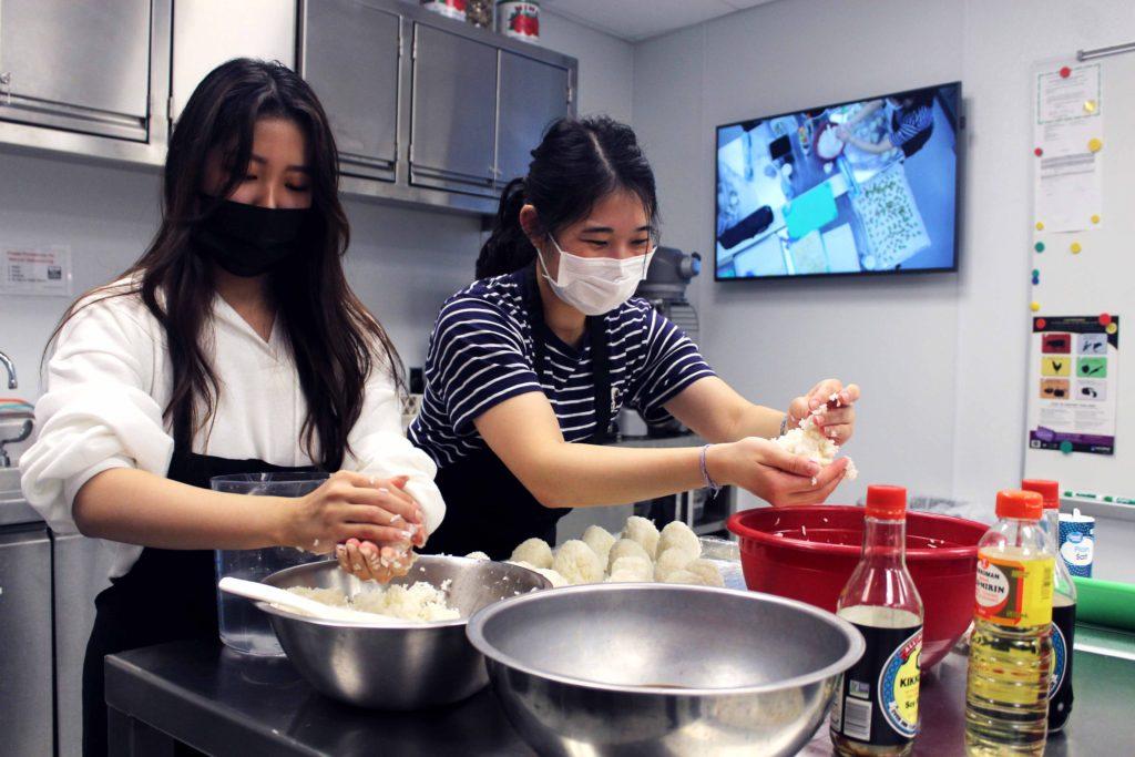 Rei Yamada `22 (left) and Sarah Amano `22 (right) make rice balls with soy sauce for Food Bazaar. In the back on the screen, you can see the image from the rotating camera in the Global Kitchen. On the cookie sheet are small cookies with vanilla, mocha and chocolate flavoring. Photo by Ariel Richards
