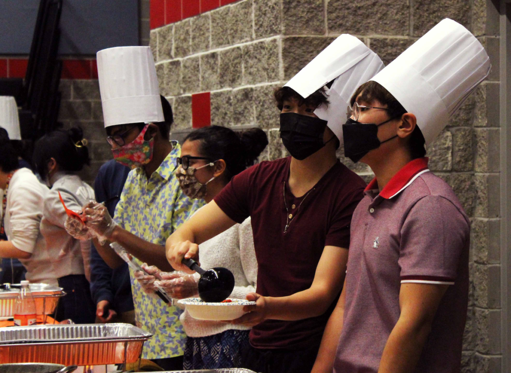 Student chefs dish out their meals while wearing adjustable hats provided by the International Student Organization. ISO ran out of hats relatively quickly, and only around half of the chefs were able to wear them. Some wore the hats more professionally than others. Photo by Maddi Shinall.