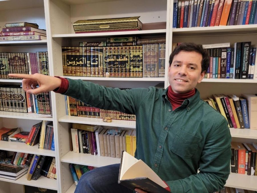 Professor Sherif Abdelkarim, English, pointing to the left as he sits with a book perched in his lap.