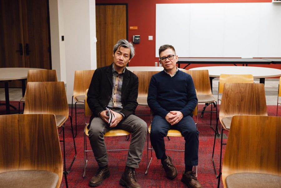 Hai-Dang Phan, on the left, and Phan Nhiên Hạo, on the right, sit in two simple wooden chairs on a red carpet looking at the camera. Their heads are tilted slightly towards each other. Their knees are spread and their hands are clasped in their laps. The chairs are part of a row and another row is visible behind them, for audience seating, but no one else is in frame. A blank whiteboard is visible on the wall behind them.