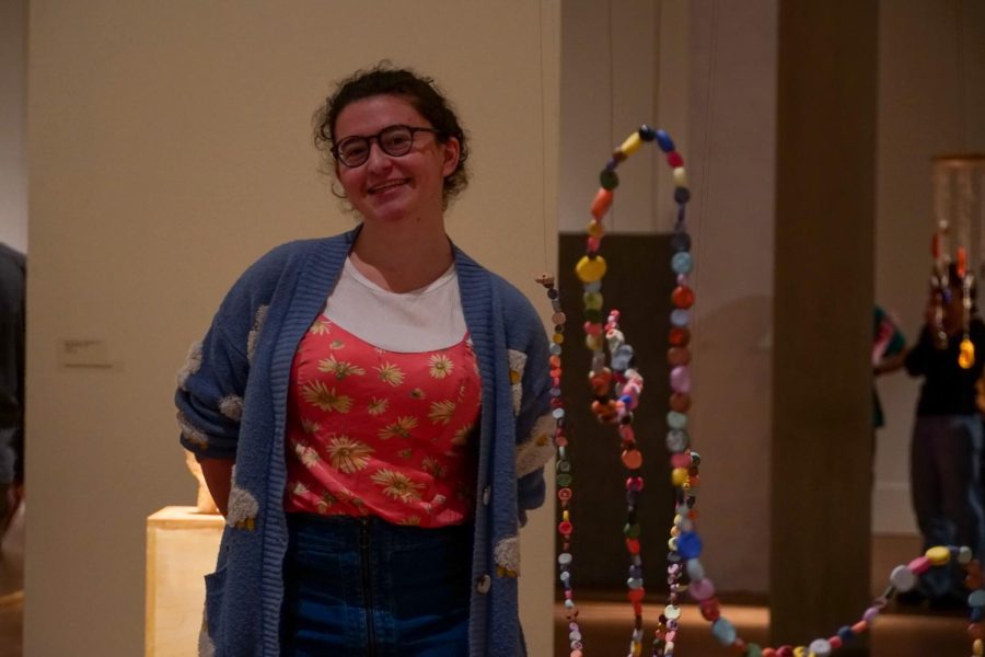 Clare standing next to her sculpture, a string of multi-colored clay beads on wire, suspended over the floor in a snake-like form.