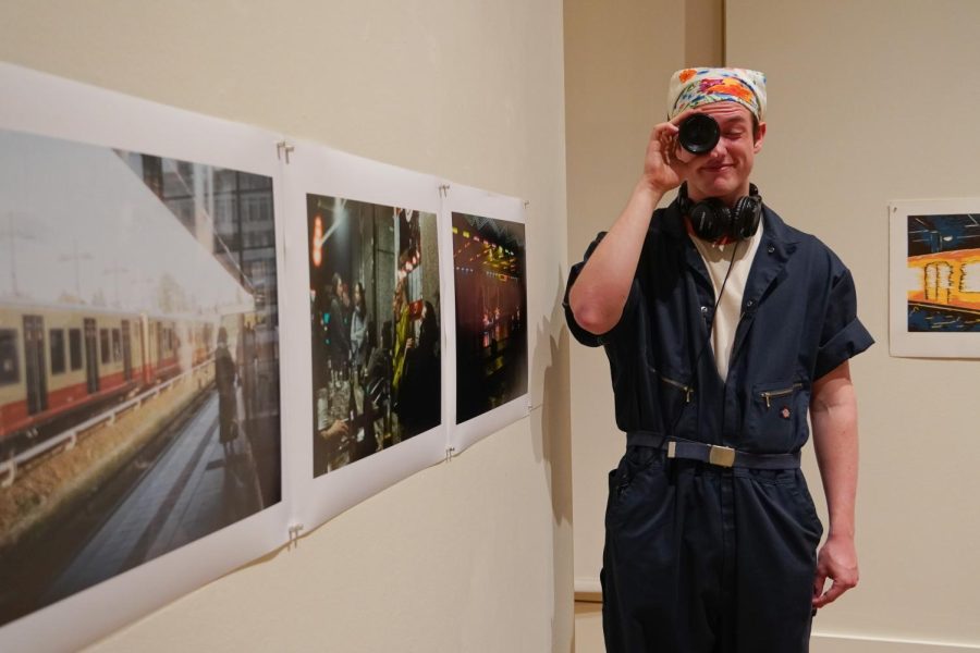 Luca standing next to his photos while holding a camera up to his eye and scrunching up his face to peer through the lens.