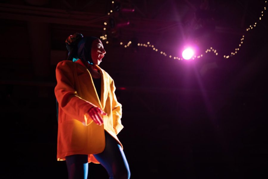 Auntie Fa striding down the stage mid-performance in a yellow blazer and vibrant head scarf.
