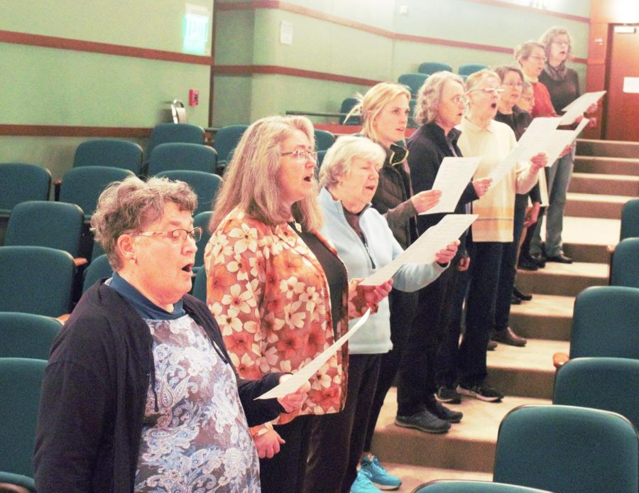Singers of various ages stand in the aisle of a theatre, each standing on a separate stair. They are singing and holding sheet music.