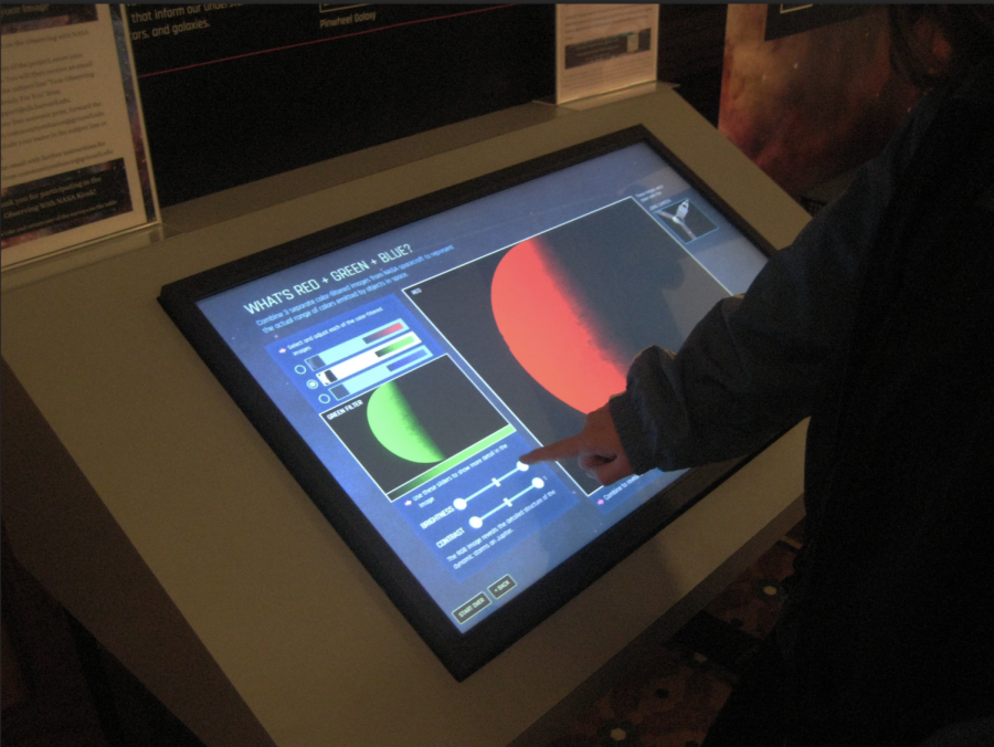 An arm with pointer finger outstretched points towards a large touchscreen displaying photos of planets and slider bars for color adjustment.