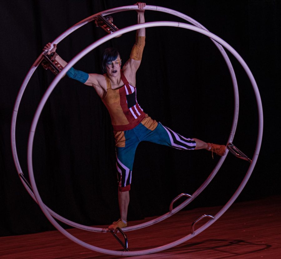 A figure in a multicolor unitard stands with her legs and arms outstretched inside one of two human-sized metal hoops that are connected with six metal spokes. It appears to be in motion. Her hands are holding onto the edge of one of the rings.