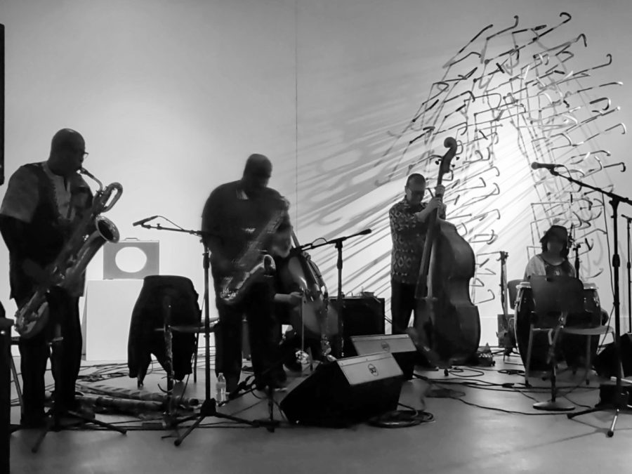 Two saxophonists, a bassist, and a drummer play music in front of a wall covered in walking canes. The image is in black and white.