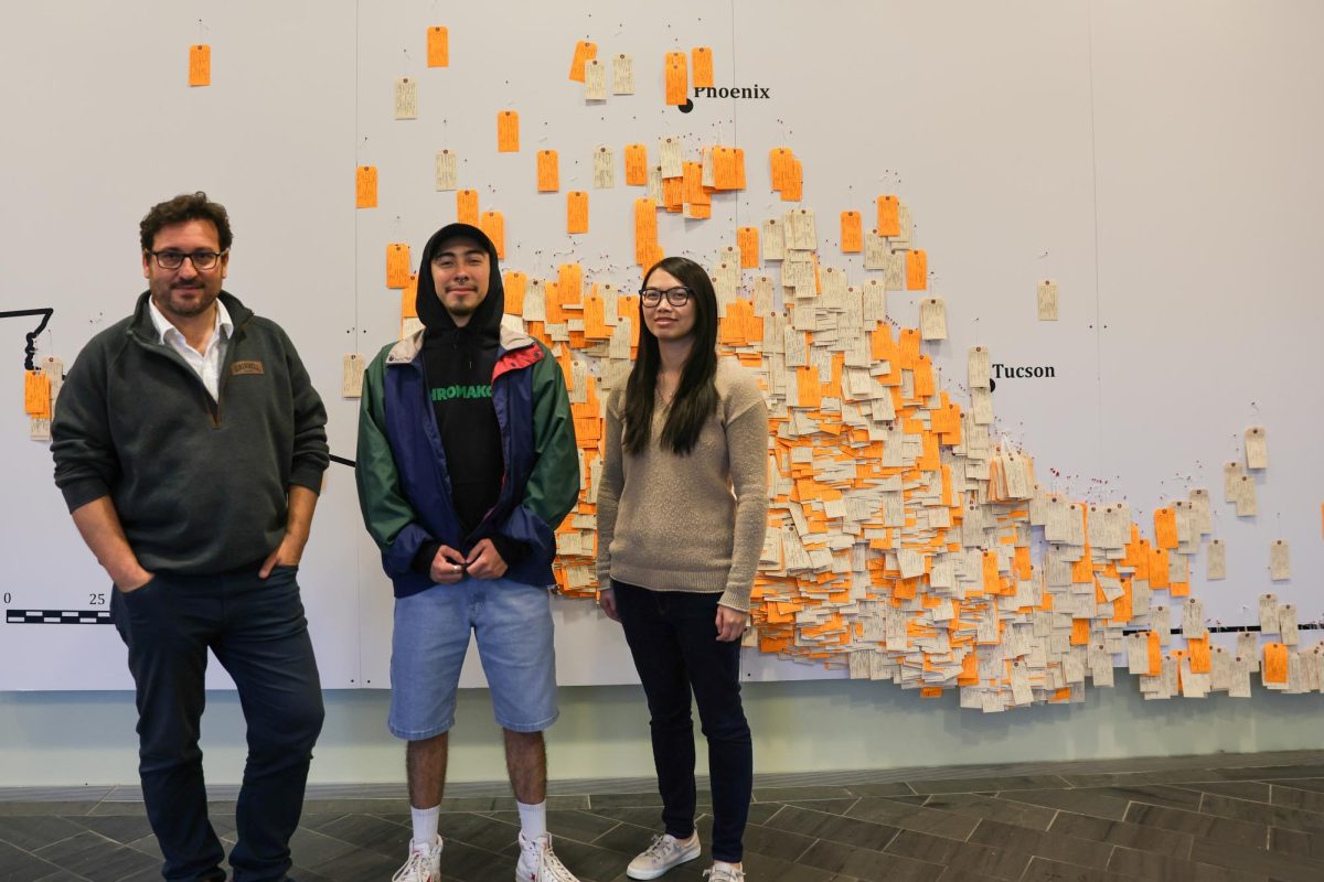 Professor Xavier Escandell (left), Jorge Salinas (middle), and Professor Laura Ng (right) stand in front of the toe tags that represent lives lost at the border in exhibit, "Hostile Terrain 94"